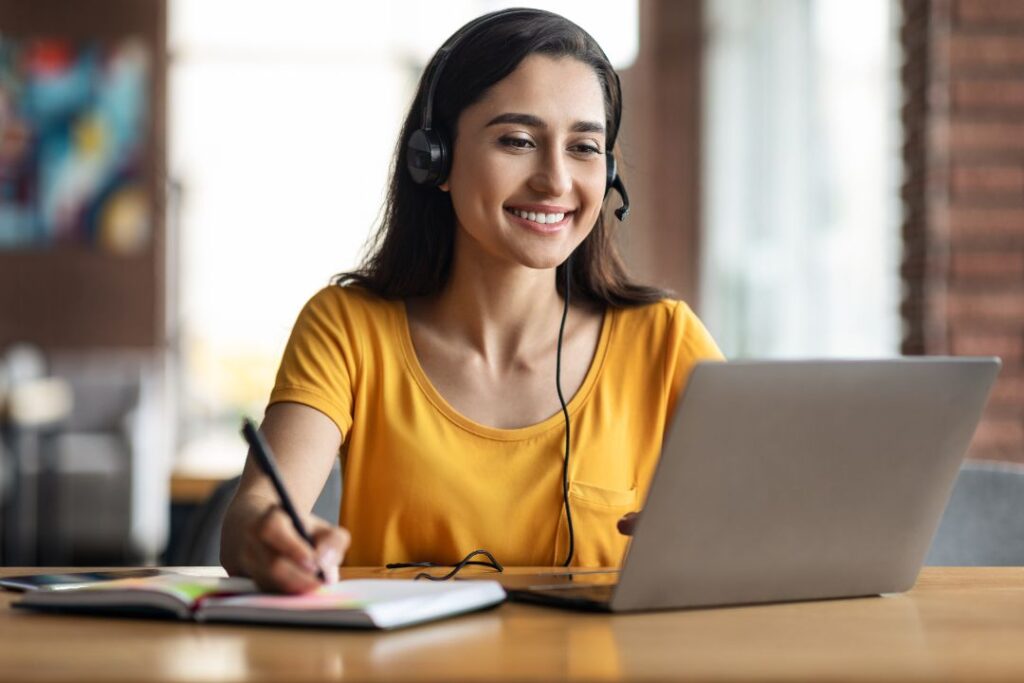 Une jeune femme souriante avec un tee-shirt jaune qui rpends des notes sur unncalepin en regardant son écran avec un casque sur les oreilles.