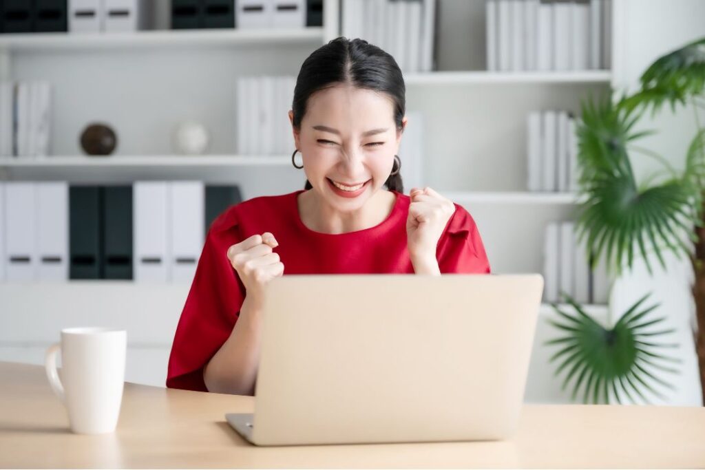 Une jeune femme de type asiatique fait un signe de victoire en souriant devant son ordinateur.